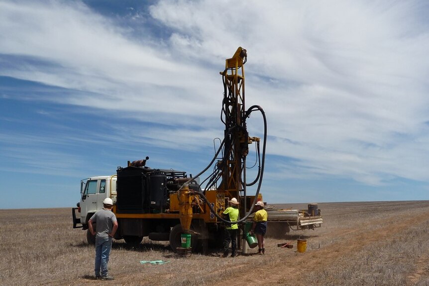 A drilling rig mounted on the back of a small truck sits in an empty field, three men are operating it.