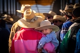 Little boy is carried by mother in her arms. Both have cowboy hats on. Boy looks backwards at camera.