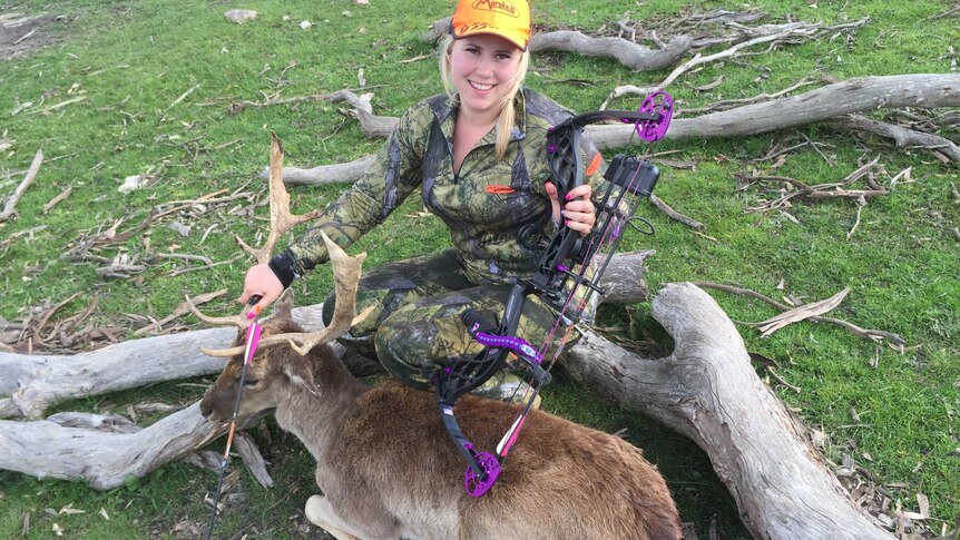 A woman with a hunting bow sitting on a log, with a dead deer in front of her.
