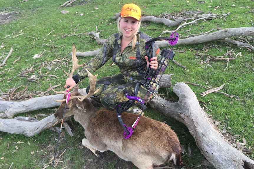 A woman with a hunting bow sitting on a log, with a dead deer in front of her.
