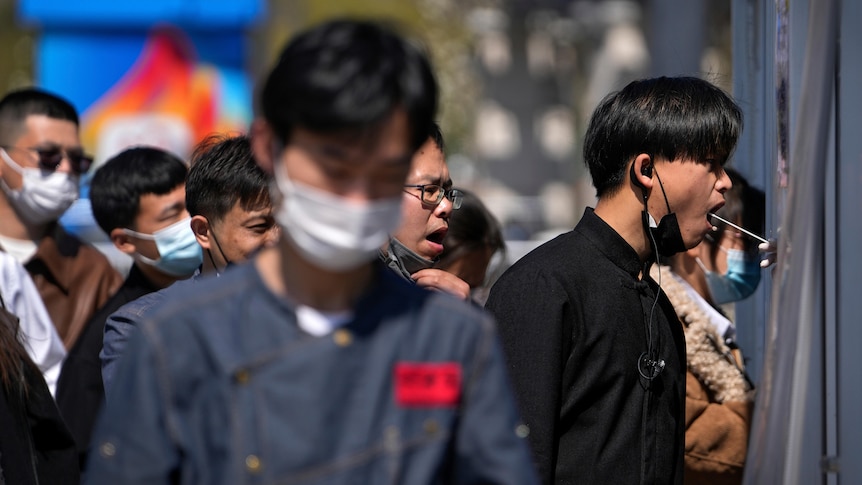 People wearing face masks line up to get their throat swabbed with a long white cotton bud.