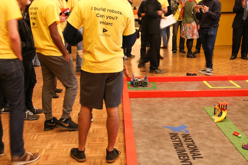 A person wearing a tshirt that says 'I build robots, can you' watches a robot car placed on the ground