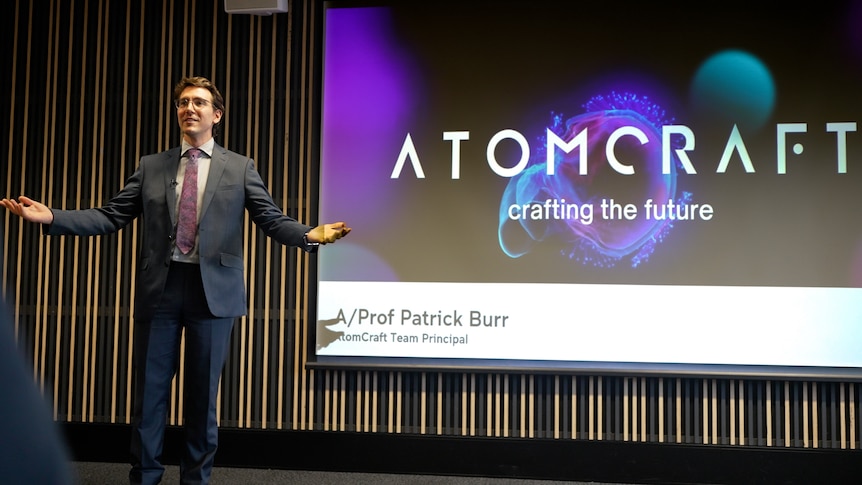 A man in a suit stands on stage, gesturing with his hands in front of a screen displaying the word "Atomcraft".