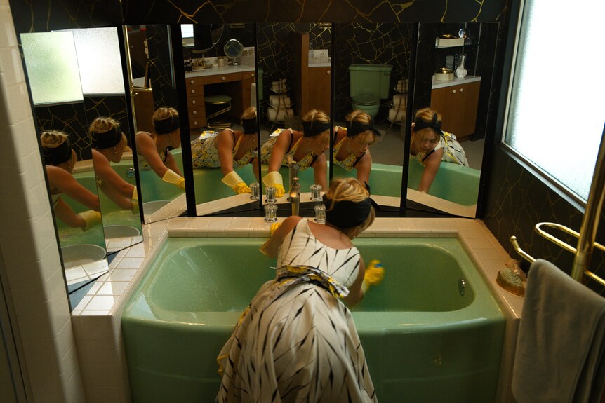 Blonde white woman in white and black 50s dress wears yellow gloves and scrubs a green bathtub.