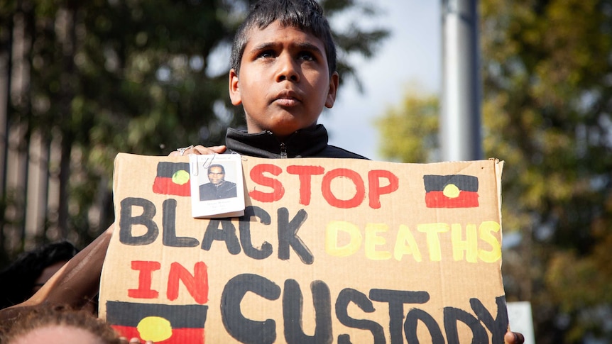 A boy holds a cardboard sign, painted with the text 'stop black deaths in custody' in the colours of the Aboriginal flag.
