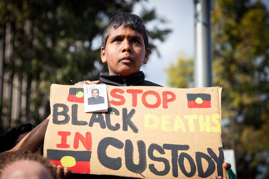 A boy holds a cardboard sign, painted with the text 'stop black deaths in custody' in the colours of the Aboriginal flag.