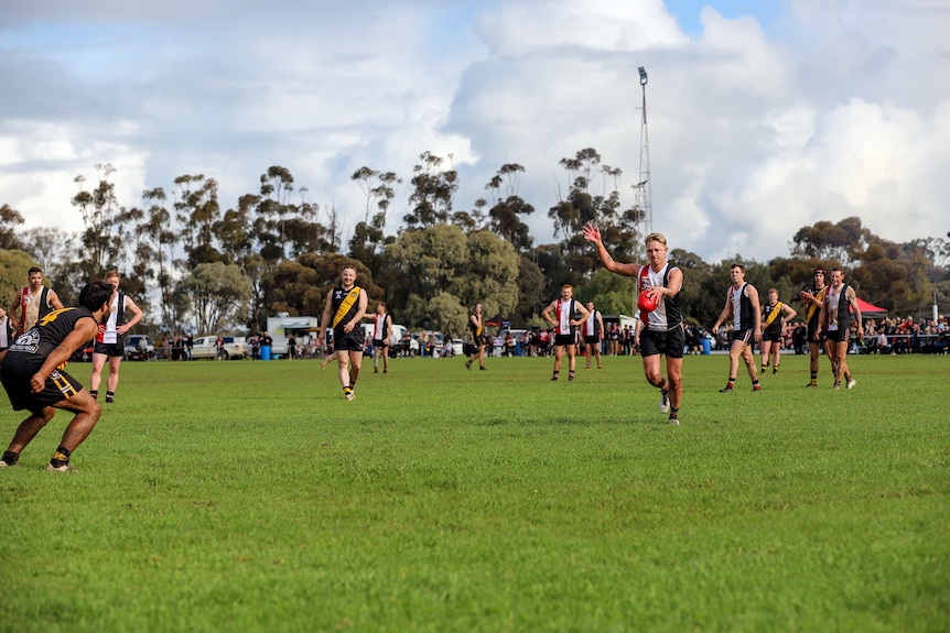 A player kicks a football over the man on the mark as other players and spectators watch on at a country footy ground 