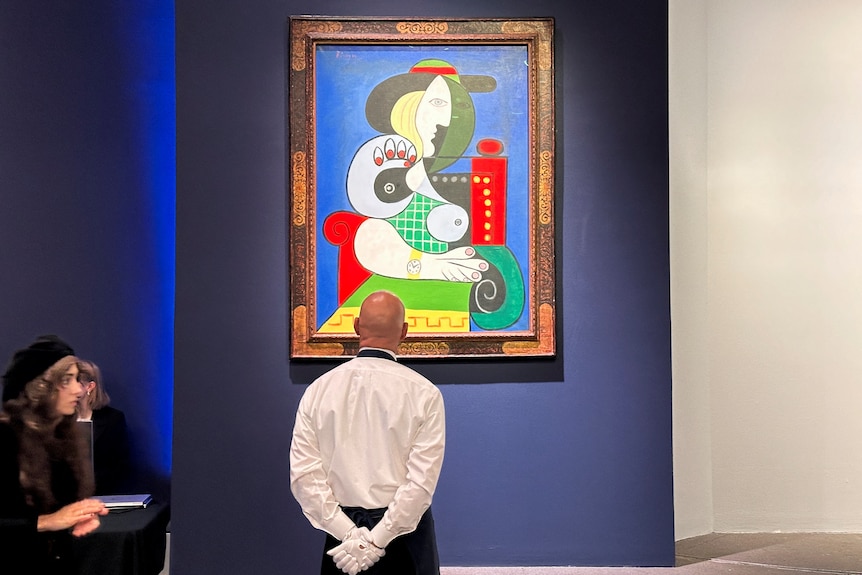 Man in white stands in front of Picasso painting with yellows and reds and blues. 