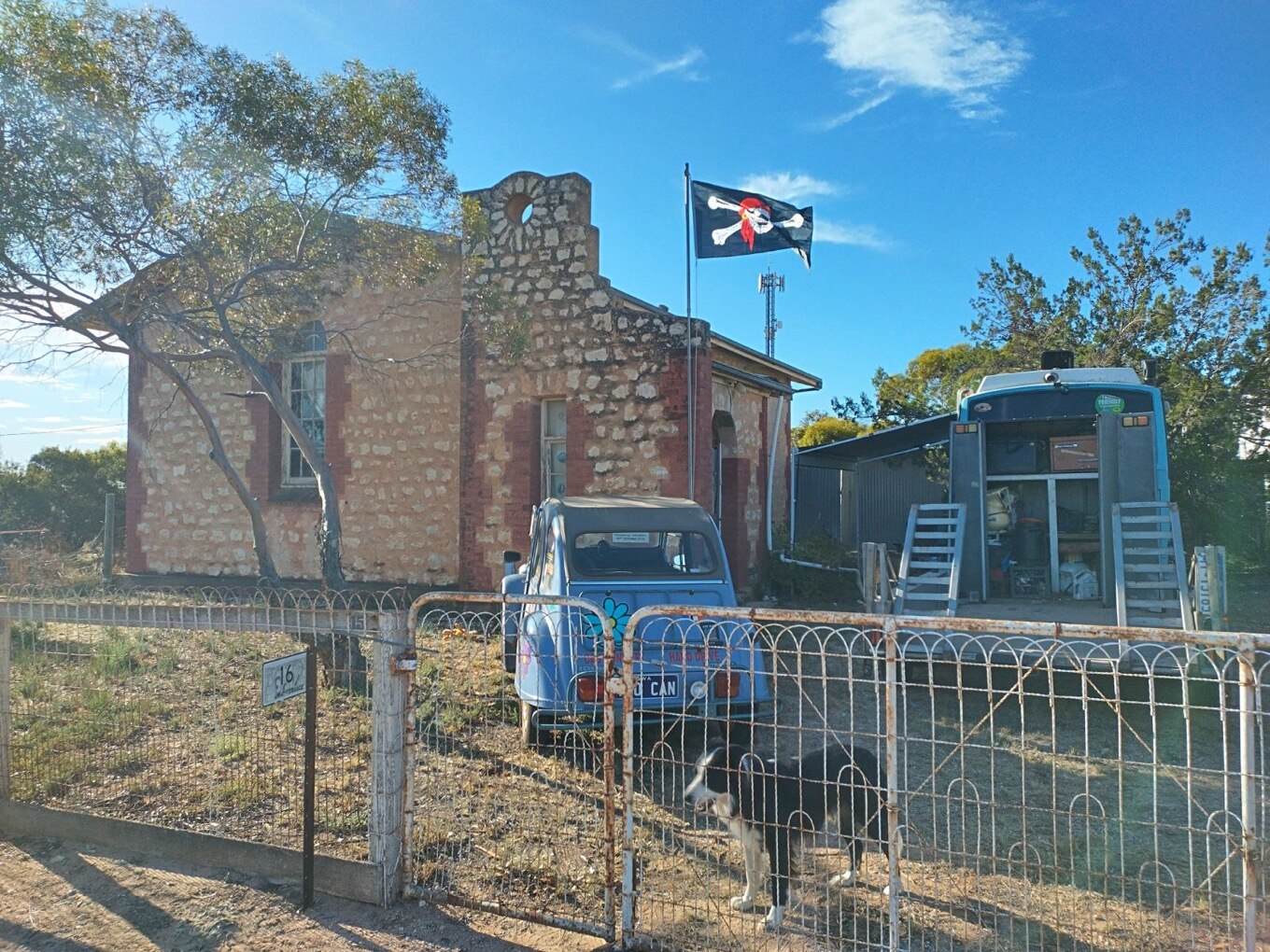 An old stone church building with a car, bus, dog and pirate flag out the front. 