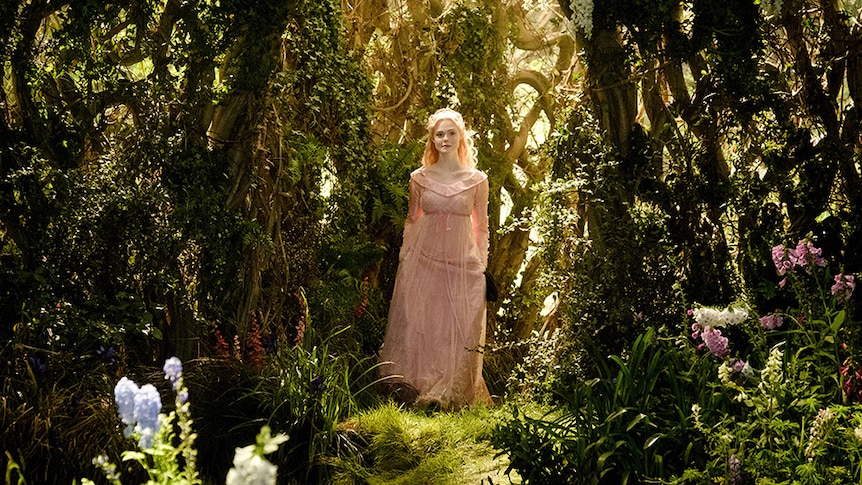 Elle Fanning wears pale pink dress and stands illuminated by warm sun light in the centre of a lush forest setting.