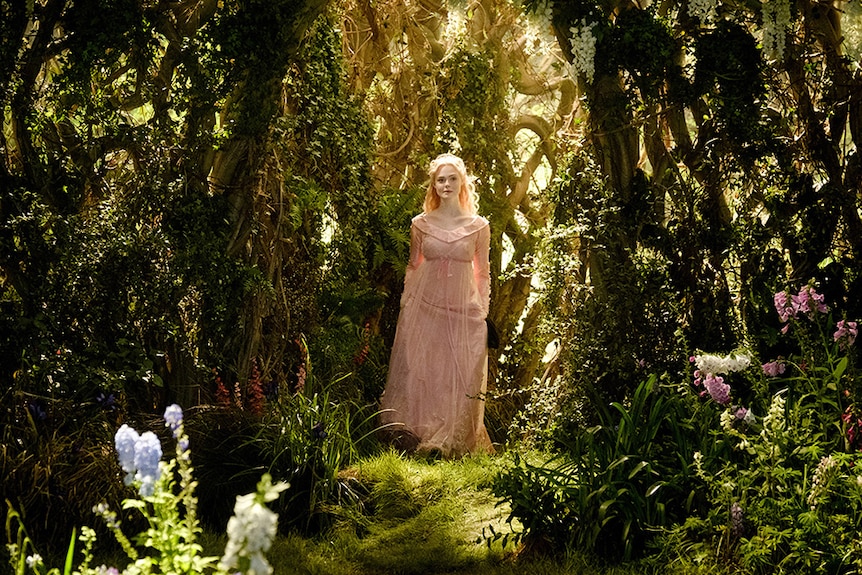 Elle Fanning wears pale pink dress and stands illuminated by warm sun light in the centre of a lush forest setting.