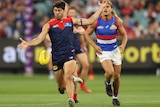 A Melbourne AFL player looks down before kicking the ball downfield, as a tired defender runs behind him. 