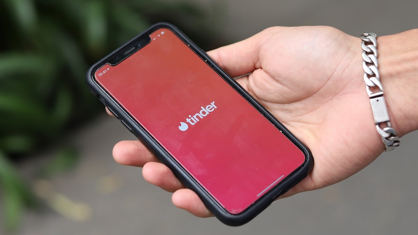 A close up of a hand with a bracelet, holding a smartphone that shows dating app Tinder. 