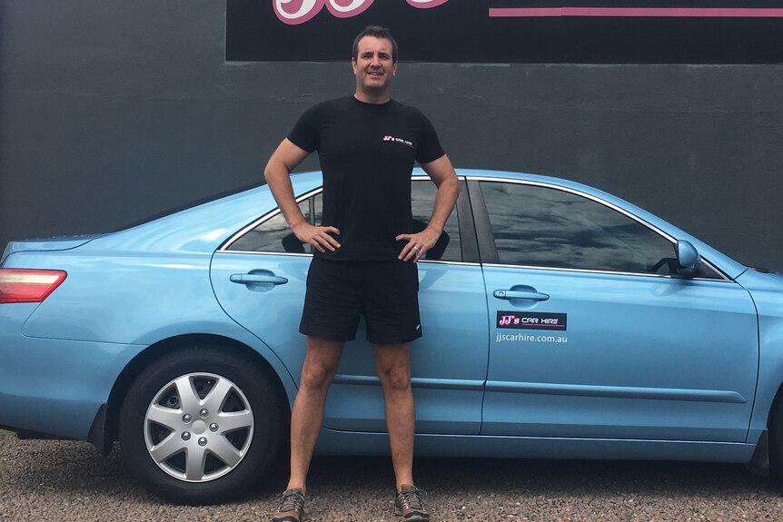 A man in a black shirt standing beside a blue sedan in front of a branded wall.