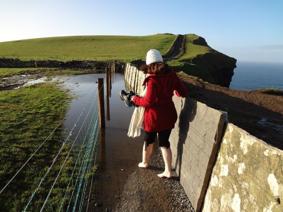 A woman walks down a stone path, wearing a red jacket and white beanie. In the distance are cliffs and the ocean.