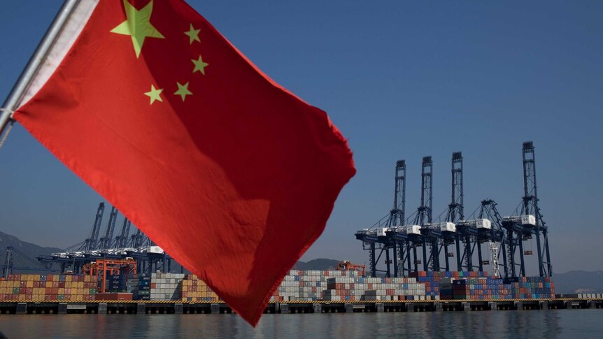 A Chinese flag attached to the back of a boat flaps in the wind as cargo containers sit on the dock of a port in China.
