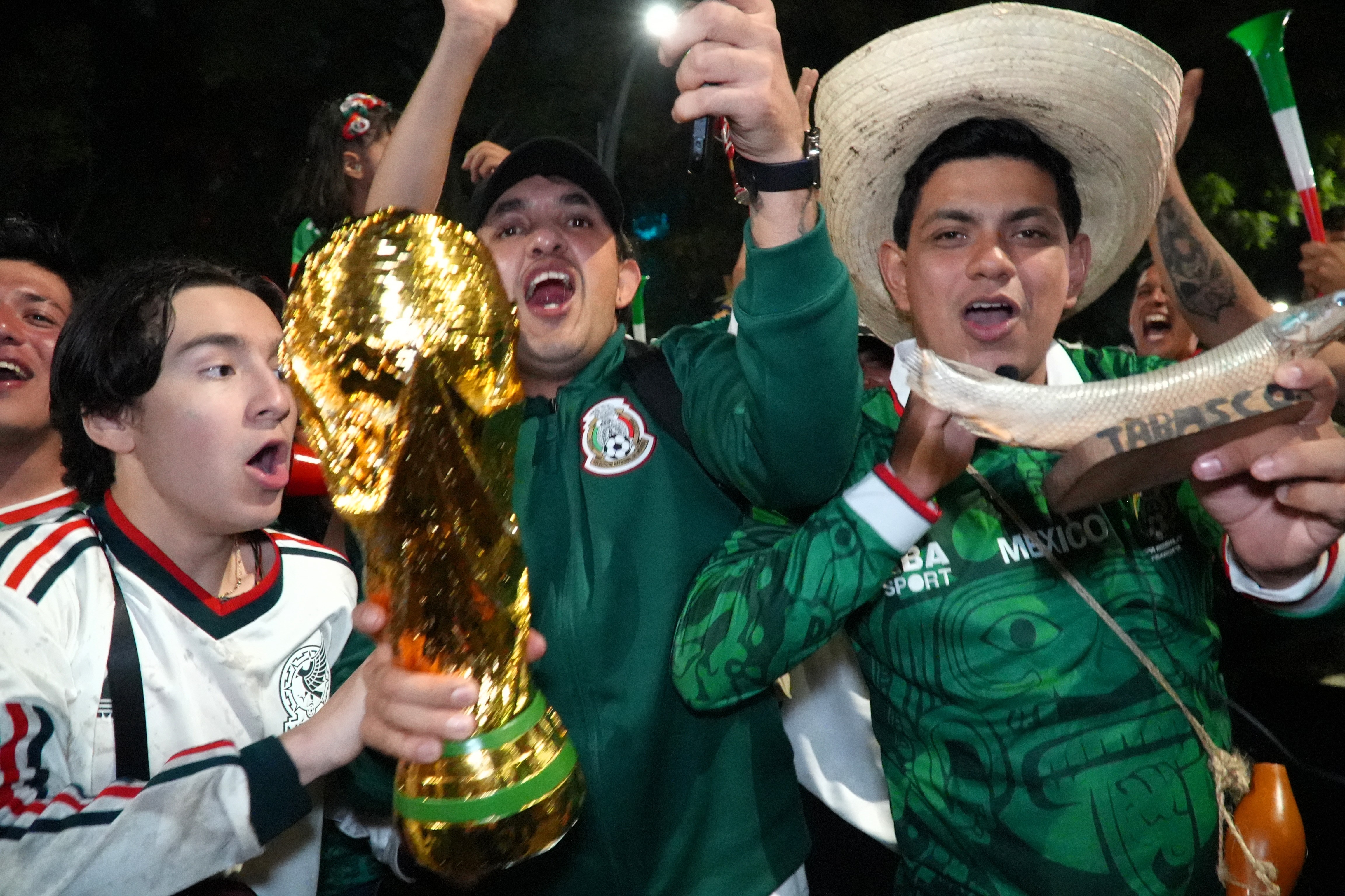 Three male Mexican football fans cheer, one is holding a World Cup trophy.