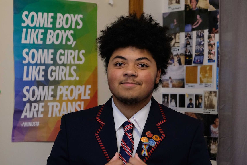 Jamil stands in his room in his school uniform. An LGBTQIA poster is in the background.