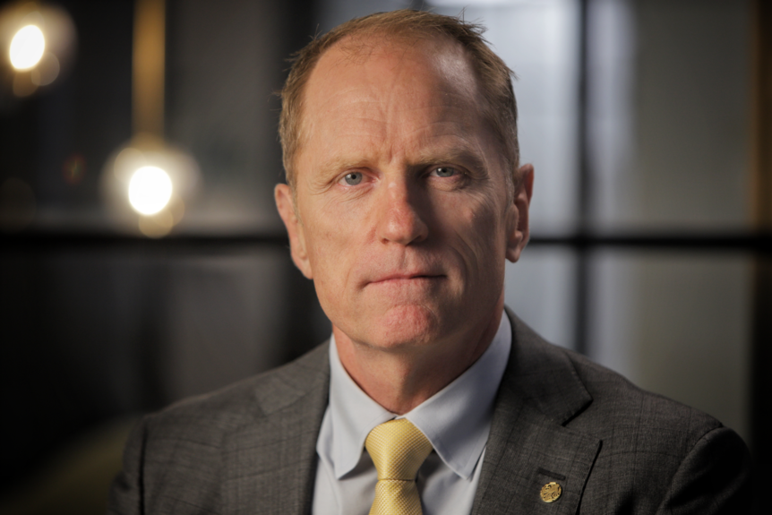 A man wearing a suit with a yellow tie, balding, looks seriously at camera.