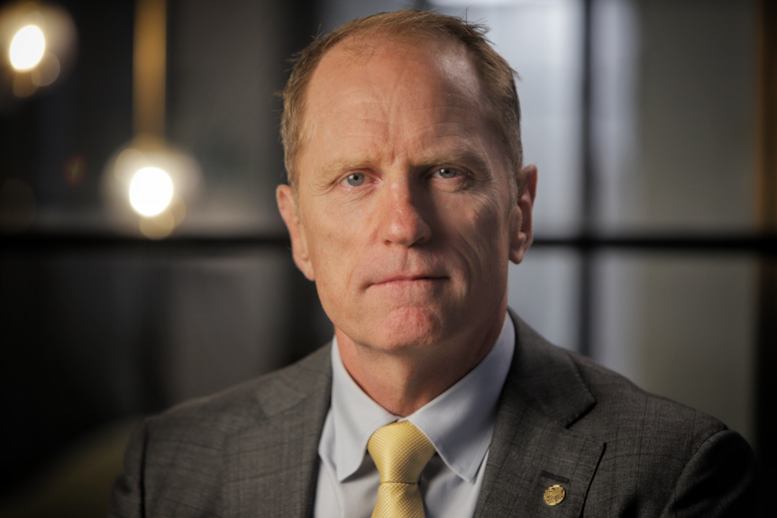 A blue-eyed, older pale man with receding red hair, wearing a grey suit jacket and shirt with a yellow tie.