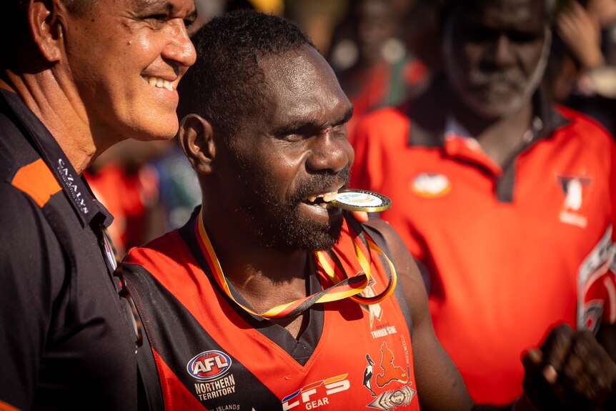 A man in a jersey holds a medal in his teeth and smiles.