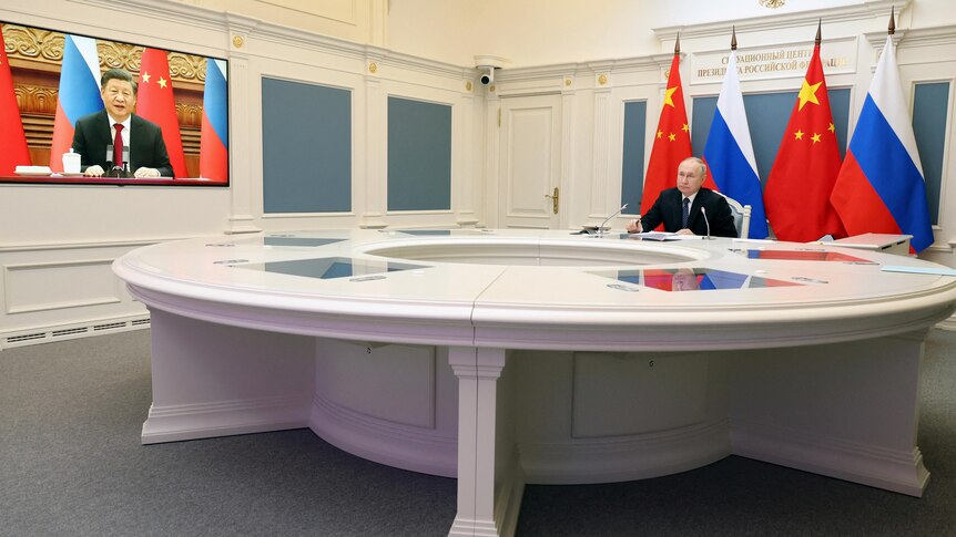Putin sits at a desk in front of Russian and Chinese flags as Xi Jinping is seen on a TV screen opposite. 