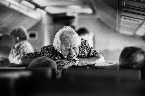 An elderly woman leans on seats on an aircraft and smiles as she talks to people.