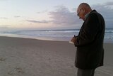 An older man, with pen and paper in hand, stands on a beach