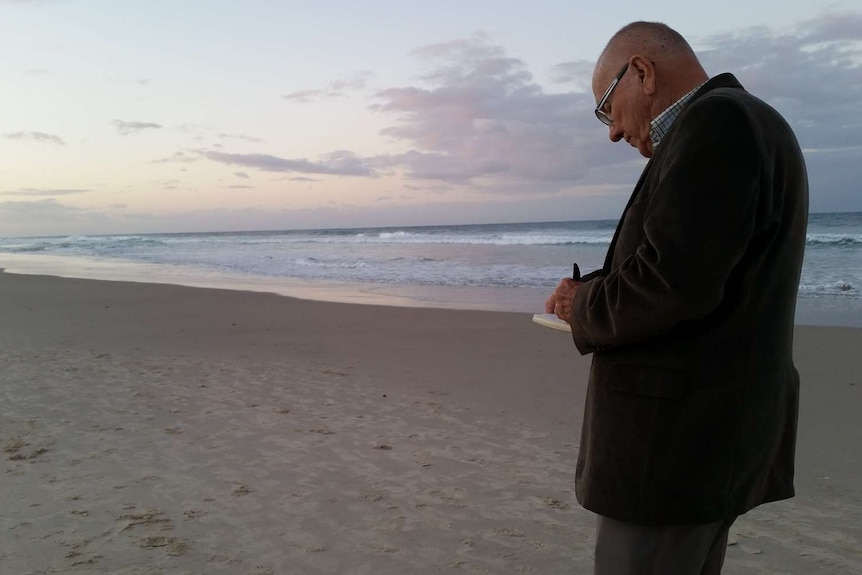 An older man, with pen and paper in hand, stands on a beach