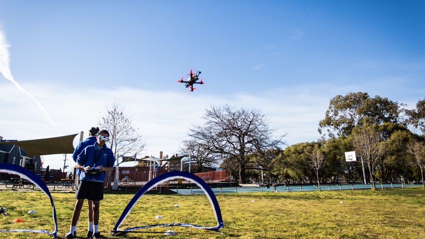 Two teenagers back-to-back drone racing on the school oval.