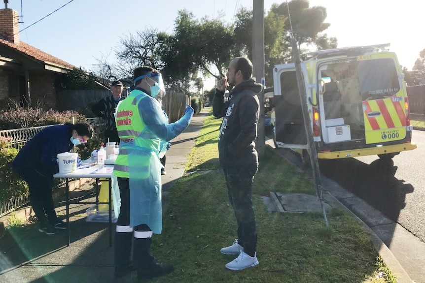 Sanjog Bisda holds a swab in his mouth in front of a coronavirus testing station and ambulance on a residential street.