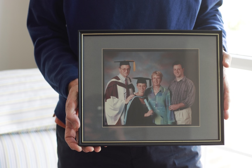 A framed photo held by hands visible either side. Photo shows two parents, adult daughter in graduation robe, and adult son.