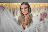 A woman holding a bridal veil with wedding dresses behind her.
