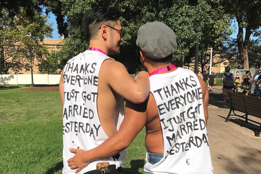 Two men wearing tank tops and showing their back, which states that they got married yesterday.