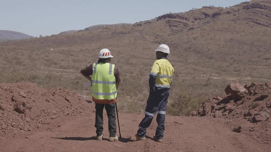 Two men in hi vis clothing and white hard hats stand looking out over the Juukan Gorge caves site.