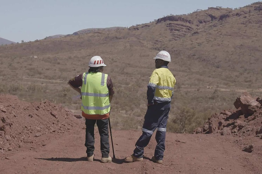 Two men in hi vis clothing and white hard hats stand looking out over the Juukan Gorge caves site.