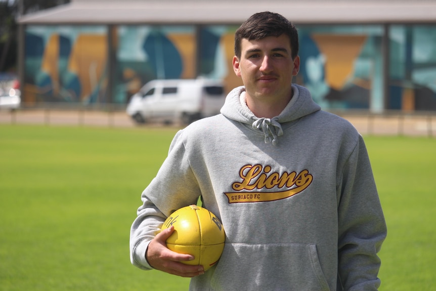 A young man stands in a hoodie on an oval holding a football