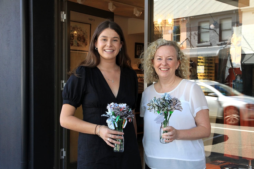 Two women holding handmade flowers in front of a shop.
