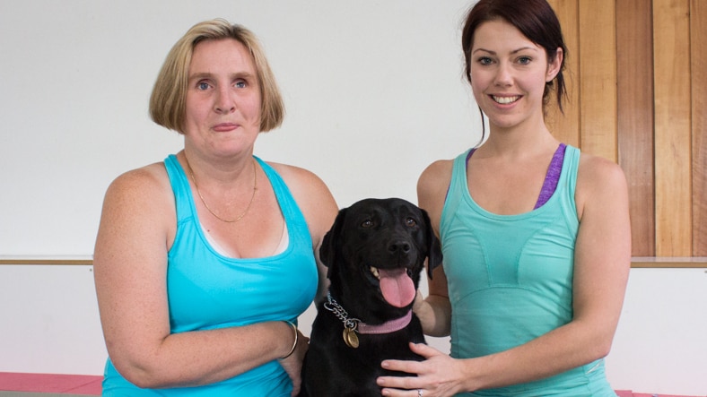 Jen Nickols sits on a floor with Kirrilee Dean. Jen's guide dog, a black labrador, sits between them.
