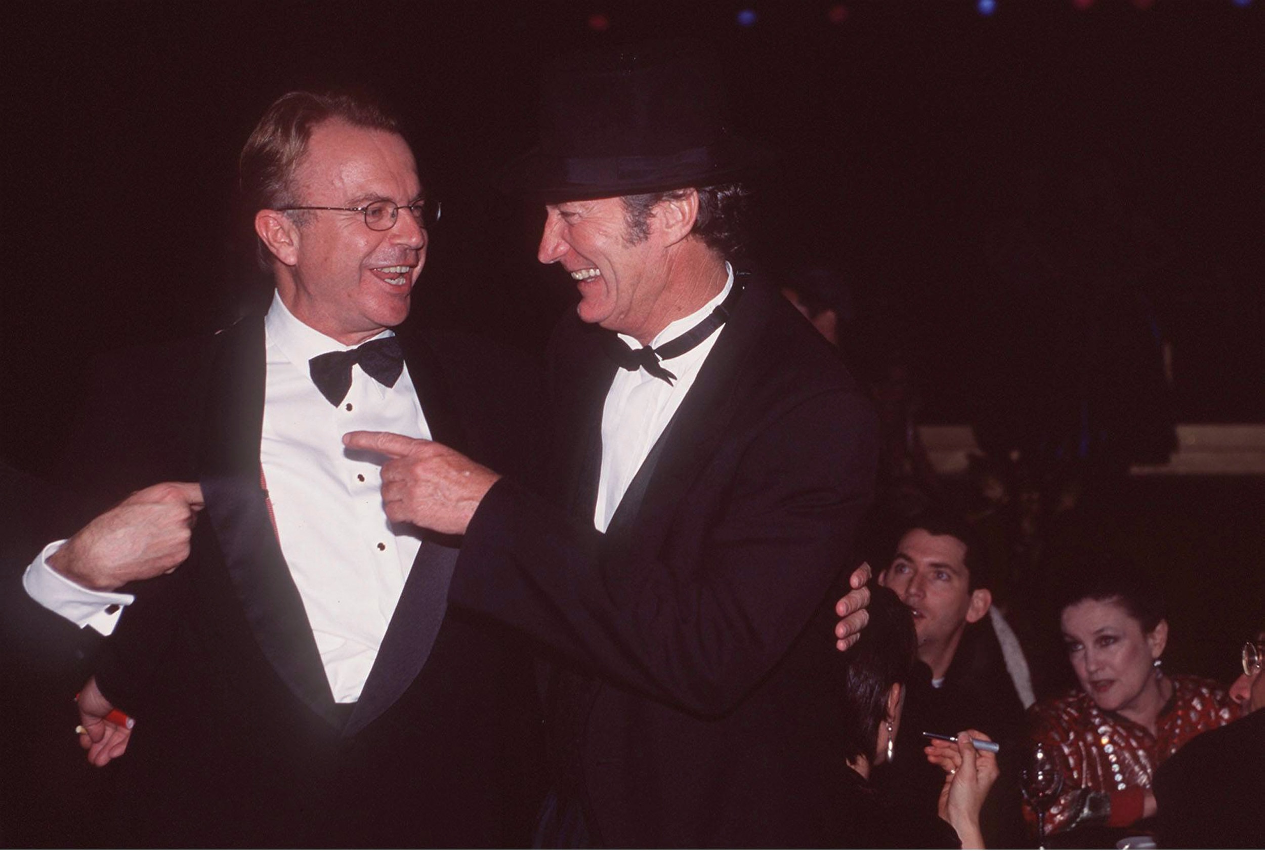 Two men dressed in black suits smile big, one pointing at the other