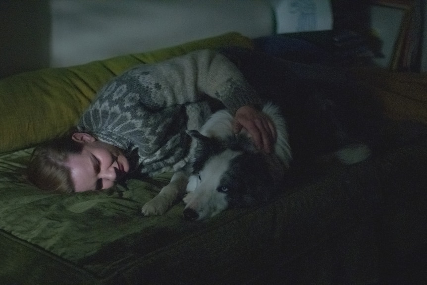 A film still of Sandra Hüller lying on a lounge in a dimly lit room, cuddling a border collie.