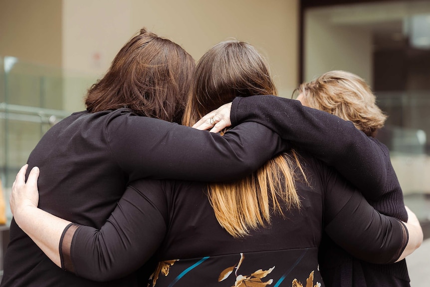 Three women dressed in black hug each other outside a building.