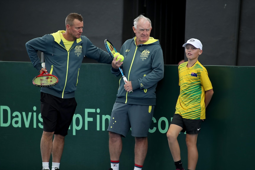 Two tennis coaches and a child stand at the back of a court during a practice session.
