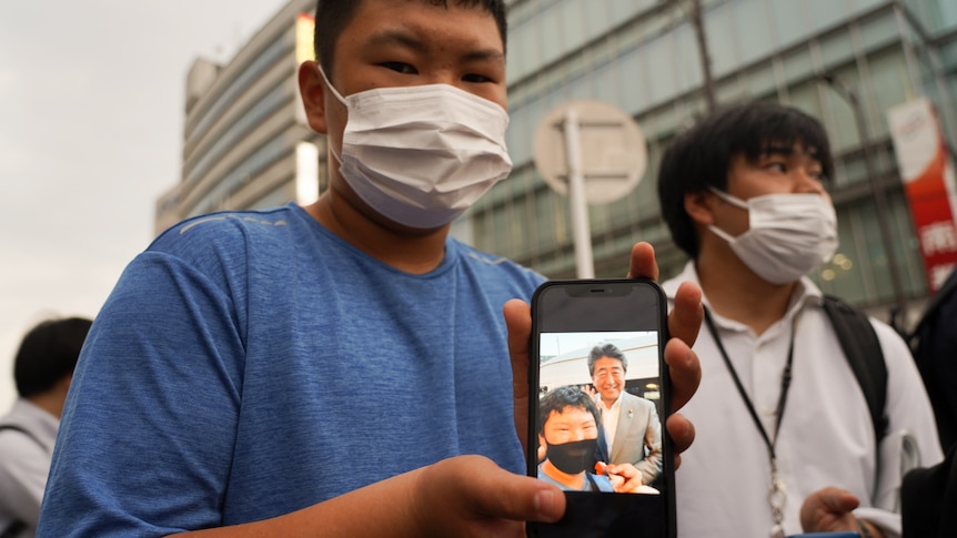 A boy wearing a blue shirt and white mask holds up an iPhone displaying a selfie of himself and Shinzo Abe