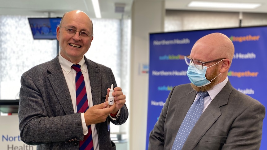 Two men, one in a surgical face mask, smiling as they hold up a small vial of a nasal spray.