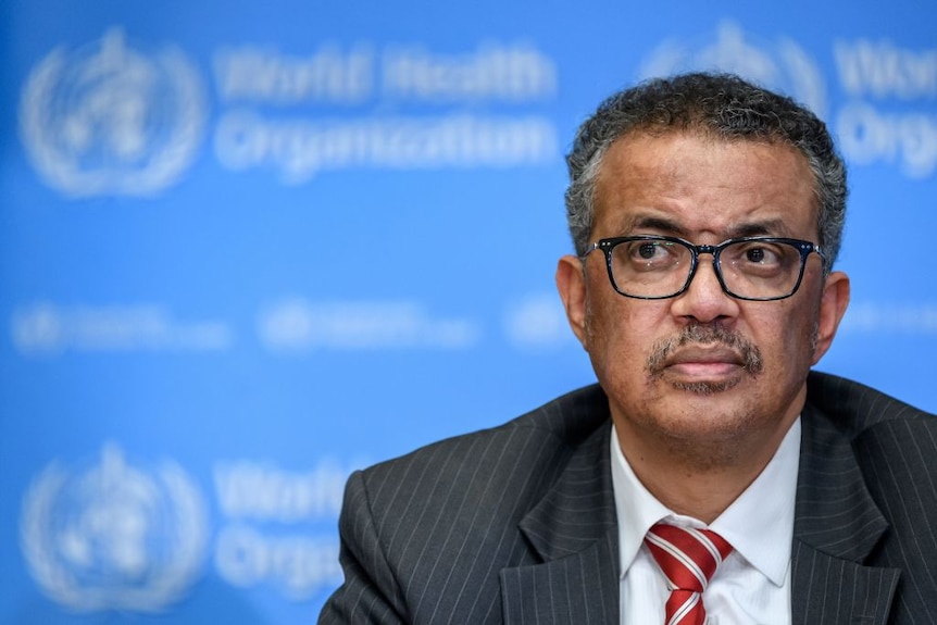 A man in a red tie and glasses suit sits in front of a blue background.