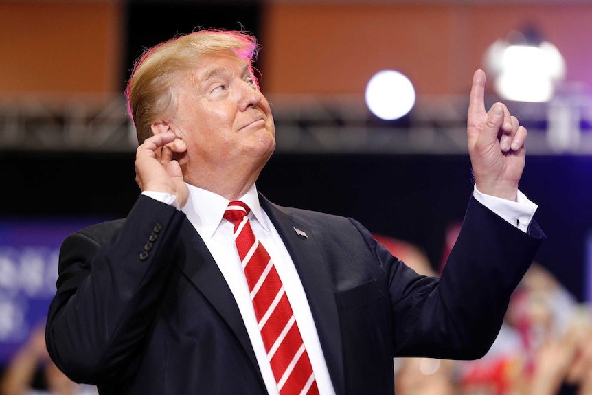 A close-up of Donald Trump with one hand on his ear and another pointing in the air