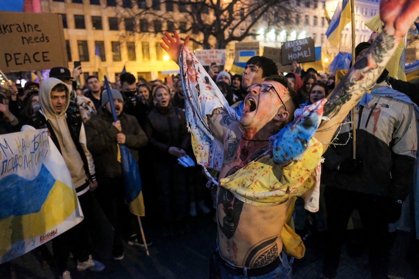 Protesters wave Ukraine flags while covered in red paint and holding signs. 