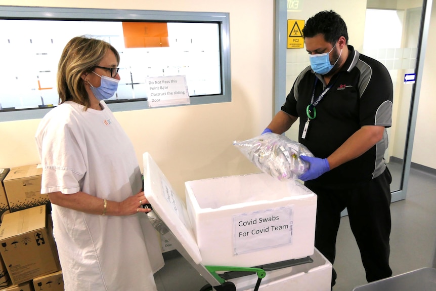 A man in surgical mask and gloves delivers COVID test swabs to a woman in a similar mask.