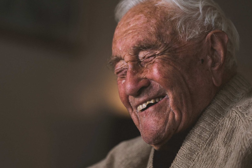 A very old man in a tan cardigan laughs against a black background.